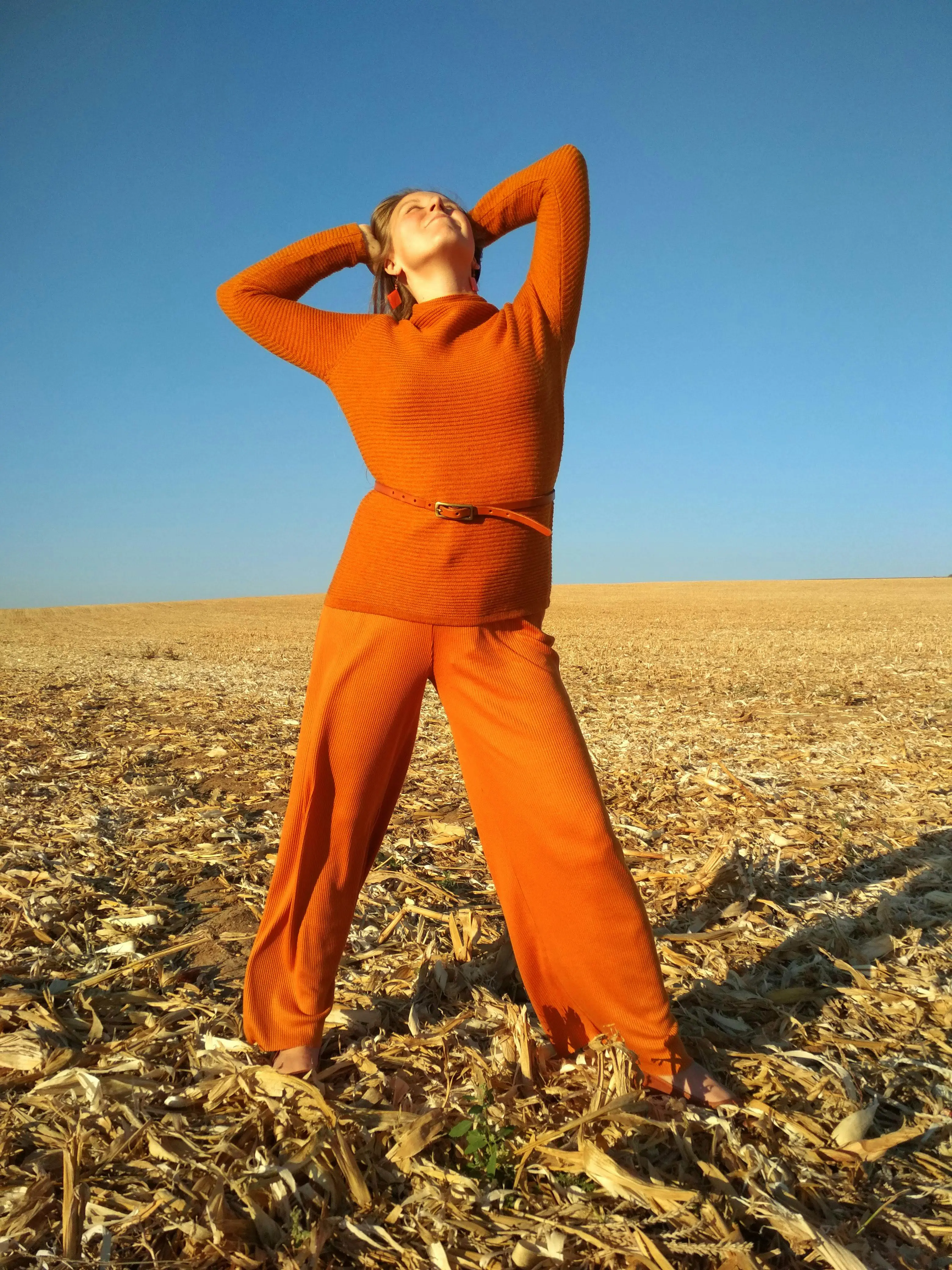 Woman wearing orange looking up, with her hands behind her head, striking a pose while standing atop a brown cornfield of short, tan dry husks, with a blue sky behind her